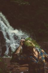 Girl with a fern leaf on her face, in the forest with a waterfall in the background