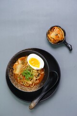 Top view of instan ramyeon or Korean noodles soup on black bowl, topped with half boiled egg, leek and Kimchi on small bowl. Isolated gray background with spoon, napkin and wooden board