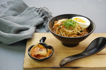 Instan ramyeon or Korean noodles soup on black bowl, topped with half boiled egg, leek and Kimchi on small bowl