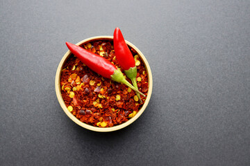 Flakes and seeds of red chili in a round bowl and two fresh peppers on a gray background.
