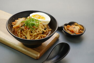 Instan ramyeon or Korean noodles soup on black bowl, topped with half boiled egg, leek and Kimchi on small bowl. Isolated gray background with spoon and wooden board