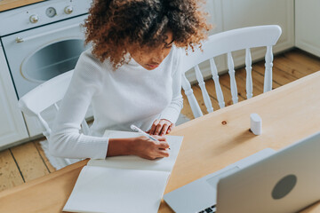 Student girl with trendy kinky hair sitting against notebook and writing notes in copybook enjoying...