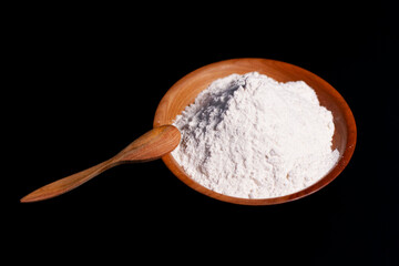 A pile of white wheat flour in a cup and on the table surface. A wooden spoon rests on the edge of the dish.