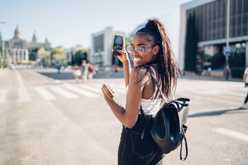 Attractive dark skinned woman in stylish wear walking on sunny day in downtown making selfie on smartphone camera, smiling african american female taking photo on mobile phone on urban settings