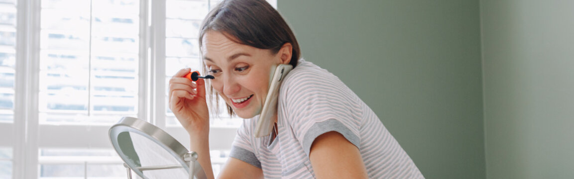 Middle age Caucasian woman applying makeup in morning and talking on phone. Young woman doing her morning routine and chatting with a friend. Web banner header.