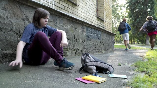 Chubby Caucasian schoolboy sitting on schoolyard ground as classmates running away. Bullied sad boy gathering books and pencils and standing up. Childhood and mockery concept.