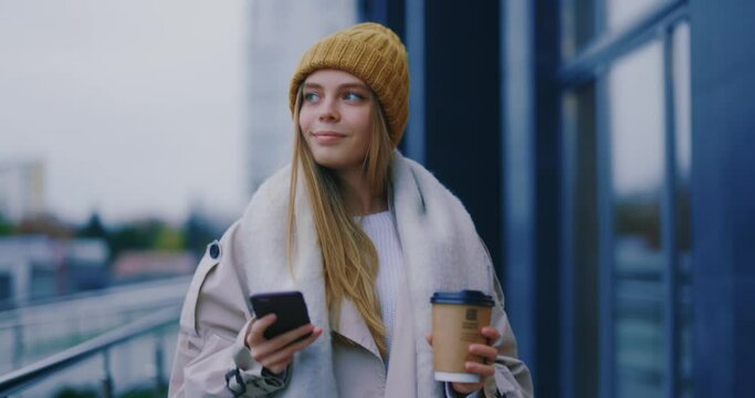 Beautiful blonde woman with coffee walking through city streets smiling enjoying autumn weather using phone for communication. Caucasian girl.