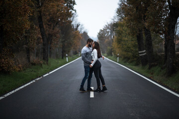 Beautiful portrait of young couple hugging while sitting on a perfect clean long road between the green forest in autumn
