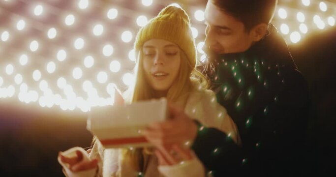 Young boyfriend making holiday surprise to his woman celebrating together giving birthday christmas present gift box hugging outdoors. Love couple. Romantic date.