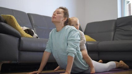 Funny kid sitting on mom's back during her home yoga class. In-house physical activity and sport together with kids