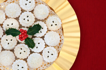 Christmas home made mince pies on a gold plate with winter holly on red tablecloth background. Festive food for the holiday season. Flat lay, top view.