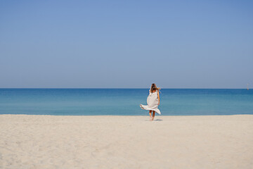 A blonde smiling girl in a maxi linen dress with fluttering hair jumping and dancing on a sand beach against blue sky and sea background.