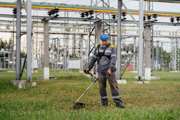 A young man mowing grass on the territory of an electric substation in overalls. Grass cleaning at the enterprise, implementation of fire safety measures