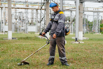 A young man mowing grass on the territory of an electric substation in overalls. Grass cleaning at the enterprise, implementation of fire safety measures