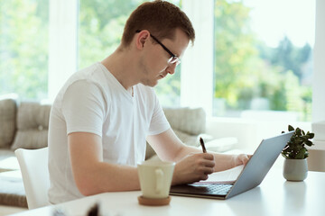 Serious young man working online with laptop at home, holding paper sitting at office desk