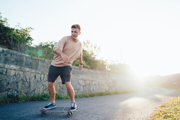 Serious caucasian 20s male skater enjoying riding on longboard on road during summer sunny vacations, handsome young hipster guy spending free time on active hobby vacations outdoors