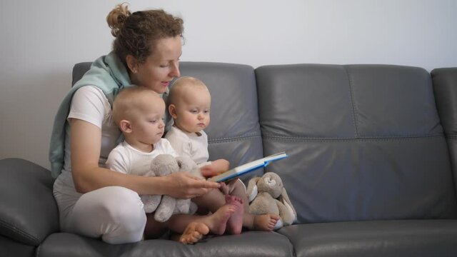 Home schooling and education for parents and children. Young mother reads book with pictures and completes exercises with two small kids on sofa