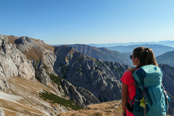 Naklejka premium A woman with big blue backpack enjoying the view on valley from top of a mountain in Hochschwab region in Austrian Alps. The flora overgrowing the slopes is turning golden. Freedom. She is happy