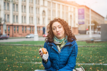 Fototapeta na wymiar Girl student in a blue jacket eats a hamburger or cheeseburger on the street