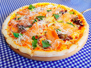 Round pizza with red fish greens and cheese stands on the table with a checkered tablecloth