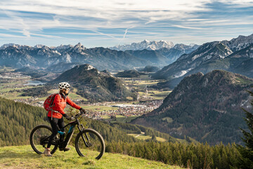 Obraz premium pretty senior woman riding her electric mountain bike in the mountains of East Allgaeu on warm autumn day with Mount Zugspitze in background