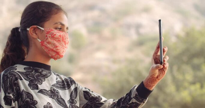 Slow-motion CU of an Indian teen girl wearing protective face mask against covid19 outdoors having a video calling communication with a technology device touchscreen handy smart pone mobile 3g network