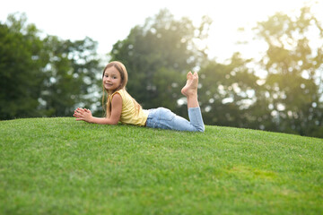 Cute little girl in casual clothes lying on the green lawn and smiling at camera while visiting park on a sunny summer day