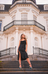 Lifestyle, blonde Caucasian in a glamorous dress and blue heels in a rustic house. Portrait of the young pretty girl on the stairs of the white house