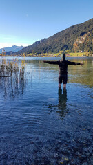 A man standing in the cold water of Weissensee, with the mountains view around. The man spreads his arms wide open in the gesture of freedom. The Austrian Alps are changing colors for winter.