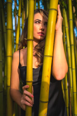 Obraz premium Lifestyle, blond Caucasian girl in a glamorous black dress in the park in nature, perched on beautiful bamboo, model looking to the right, vertical photo
