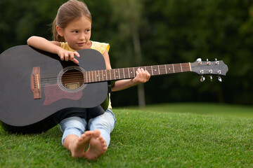 Adorable little girl looking aside and holding guitar sitting on a green grass in park, spending...