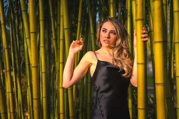 Fototapeta na wymiar Lifestyle, blonde Caucasian girl in a black strappy dress, in beautiful bamboo in the park outdoors, the girl perched holding on to the pole and looking to the left