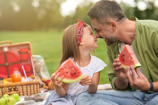 Adorable little girl playing with her loving daddy while eating watermelon, family having a picnic in the green park on a summer day