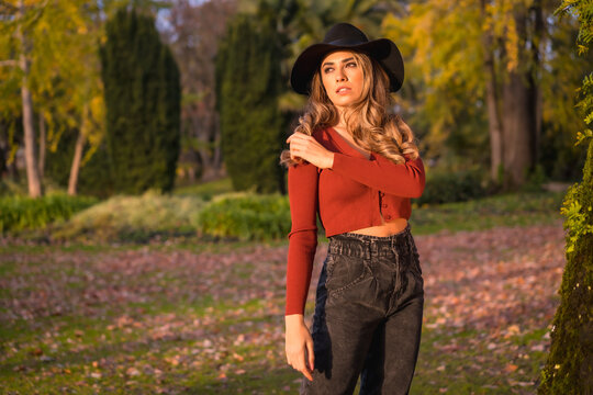 Lifestyle, blonde Caucasian girl in a red sweater and black hat, enjoying nature in a park with trees, portrait of the young woman enjoying the autumn sun