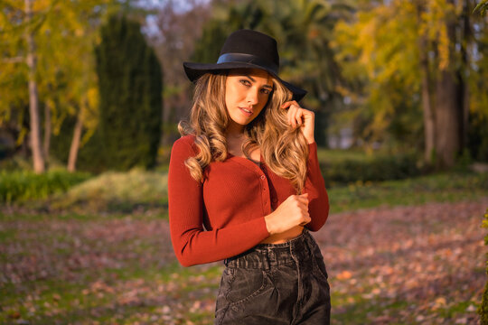 Lifestyle, blonde Caucasian girl in a red sweater and black hat, enjoying nature in a park with trees, portrait of the young woman enjoying the autumn sun