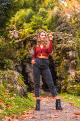 Autumn lifestyle, blonde Caucasian girl in a red sweater and jeans and with high heels, enjoying nature in a park with a natural cave in the background