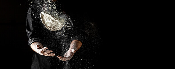 flying pizza dough with flour scattering in a freeze motion of a cloud of flour midair on black. Cook hands kneading dough. copy space - obrazy, fototapety, plakaty