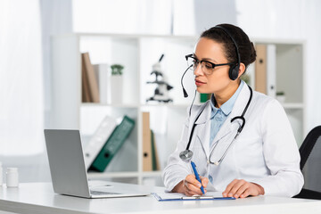 Fototapeta na wymiar African american doctor in headset writing on clipboard, while looking at laptop during online consultation on blurred background