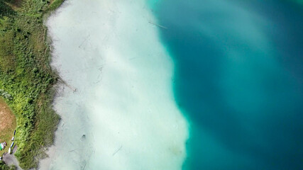 A top down, drone capture of Weissensee lake in Austrian Alps. The water changes color from white, turquoise to navy blue. Green shore of the lake. A bit of overcast. Serenity and peacefulness