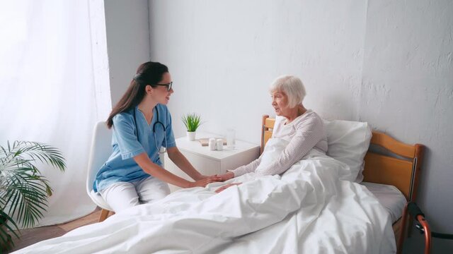 elderly woman sitting in bed and talking to brunette geriatric nurse