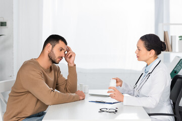 African american woman holding bottle with pills while looking at thoughtful patient on blurred background