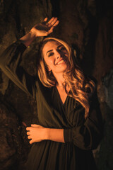 Lifestyle, a young Caucasian blonde in a black dress in a photo inside a cave, illuminated with yellow light. Very seductive pose of the model