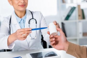 Cropped view of african american doctor pointing with pen at bottle with pills in hand of patient on blurred background