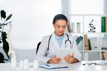 Positive african american doctor holding digital tablet, while sitting at workplace on blurred background