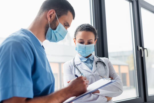 African American Doctor In Medical Mask Standing Near Nurse Writing On Clipboard On Blurred Foreground