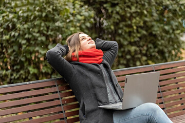 Fototapeta premium A young woman, with her hands behind her head, feels peace, rests after work, sits on a wooden bench in the park a laptop on her knees. Break during work. Preventing stress and relieving anxiety.