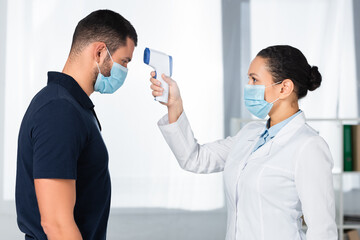 African american doctor in medical mask using infrared thermometer on patient in clinic