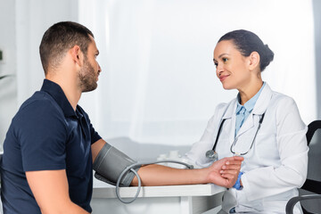 Fototapeta premium Smiling african american doctor measuring blood pressure of adult patient, while sitting at workplace
