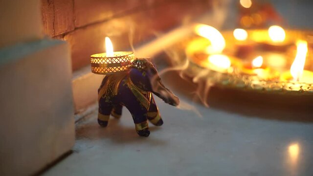 Hand moving in to pick an elephant incense holder with smoke coming out of it and beautiful gold painted earthenware diya lamps filled with oil and lit with a flame in the background as decor for the