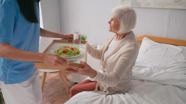 young nurse carrying tray with breakfast to aged patient